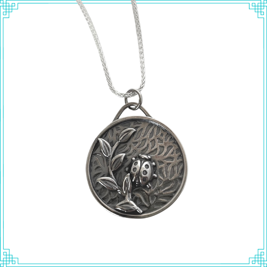 Sleeping Fox handmade silver jewelry created this round pendant with a lady bug and leafy branch elements. The background is a bed of leaves. This pendant hangs from an 18" wheat chain.