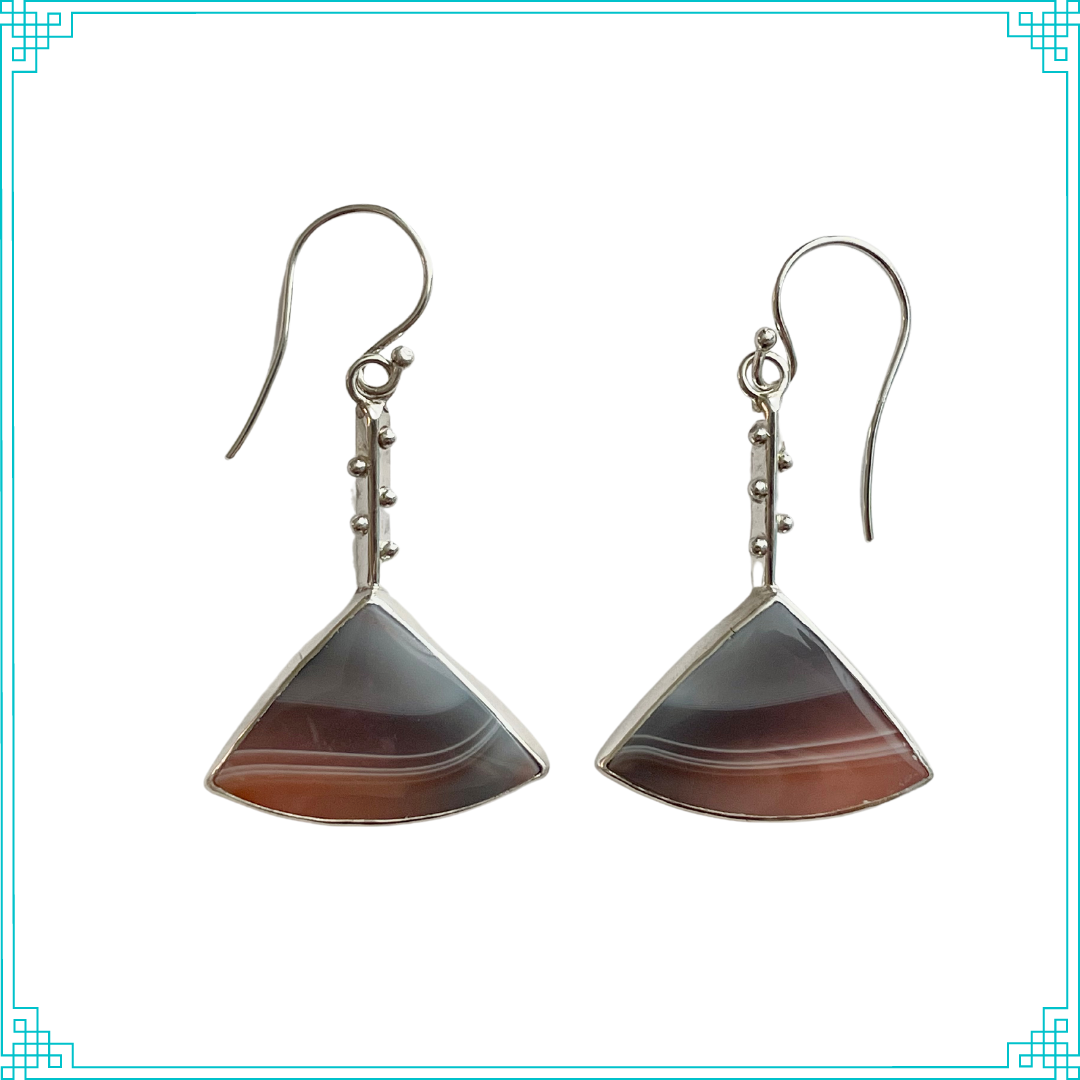 These Sleeping Fox banded agate earrings have a soft pallet with smokey blue lines fading into a hazy light peach color. Truly remarkable! The stones hang from a triangle wire studded with sterling silver balls. On the back, the hallmark sits upon distinctive curved lines resemble tree rings. We hand make the sterling ear wires.