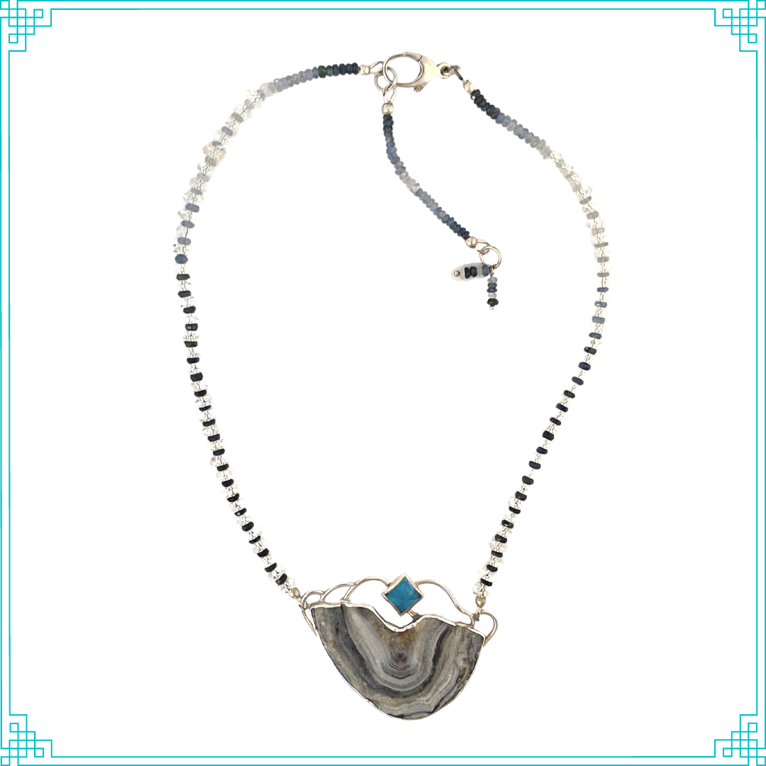 The chain features color graduating faceted shades of blue sapphires gleaming through faceted Herkimer diamond beads. The chain is attached to the pendent, and can be worn at two lengths (16in and 18in). The pendent is made from sterling silver, displaying a pyramid Swiss Blue Topaz. The open back bezel around the Topaz allows light to shine through. Below, is a Brazilian agate with a shimmery druzy surface. The blue hues and meandering druzy channels in the stone give this piece its name.
