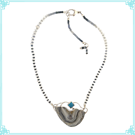 The chain features color graduating faceted shades of blue sapphires gleaming through faceted Herkimer diamond beads. The chain is attached to the pendent, and can be worn at two lengths (16in and 18in). The pendent is made from sterling silver, displaying a pyramid Swiss Blue Topaz. The open back bezel around the Topaz allows light to shine through. Below, is a Brazilian agate with a shimmery druzy surface. The blue hues and meandering druzy channels in the stone give this piece its name.