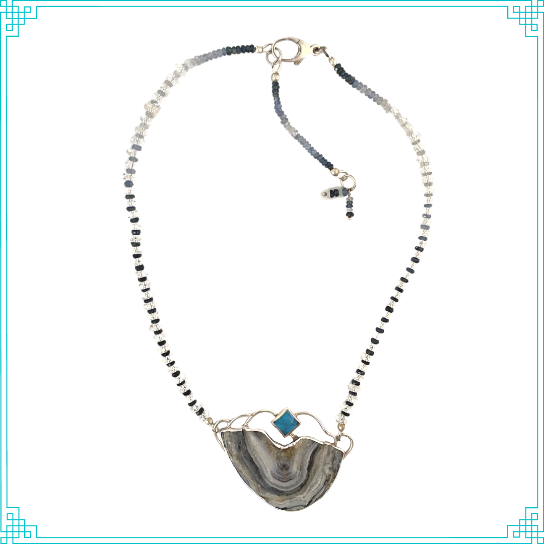 The chain features color graduating faceted shades of blue sapphires gleaming through faceted Herkimer diamond beads. The chain is attached to the pendent, and can be worn at two lengths (16in and 18in). The pendent is made from sterling silver, displaying a pyramid Swiss Blue Topaz. The open back bezel around the Topaz allows light to shine through. Below, is a Brazilian agate with a shimmery druzy surface. The blue hues and meandering druzy channels in the stone give this piece its name. 
